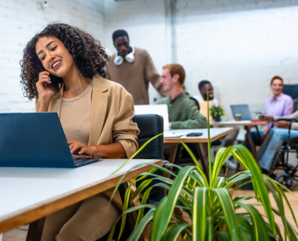 Woman talking to the mobile phone and using laptop in a coworking space with coworkers on the background
